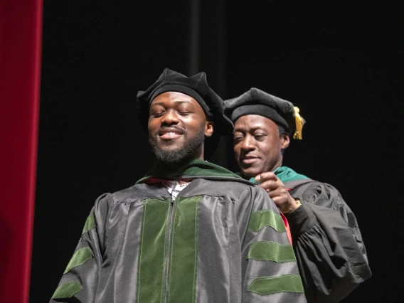 A University of Arizona College of Medicine – Tucson student dressed in a graduation cap and gown has a ceremonial hood placed over his shoulders by his father, who is also dressed in graduation regalia.