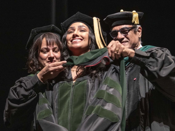 Two University of Arizona College of Medicine – Tucson professors place a ceremonial graduation hood over the shoulders of their daughter. All are dressed in caps and gowns and smiling. 