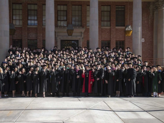 More than 100 University of Arizona College of Medicine – Tucson class of 2024 graduates and faculty stand on the stairs outside a building on the UArizona campus. All are dressed in graduation regalia. 