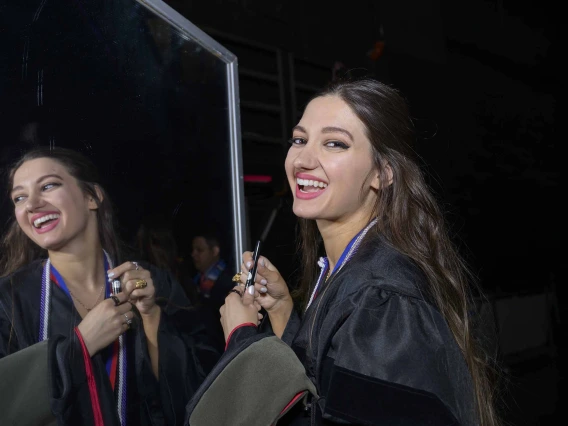 A University of Arizona R. Ken Coit College of Pharmacy student wearing graduation regalia smiles as her image is reflected in a mirror. 