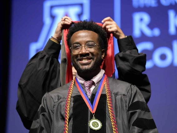 A University of Arizona R. Ken Coit College of Pharmacy student in a graduation gown smiles as a professor places a graduation hood over his head. 