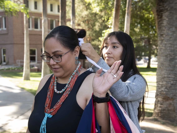 A University of Arizona Mel and Enid Zuckerman College of Public Health student wearing a large beaded necklace stands outside as her niece ties her hair in a bun.