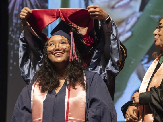 A University of Arizona Mel and Enid Zuckerman College of Public Health student smiles as she has a hood placed over her shoulders by a faculty member. 