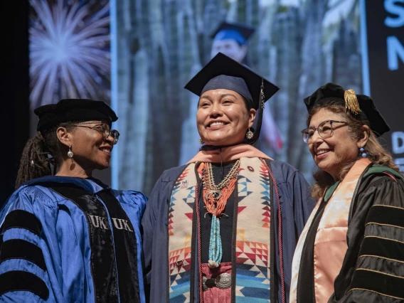 A University of Arizona Mel and Enid Zuckerman College of Public Health student smiles as she stands between two faculty members. All are wearing graduation regalia.