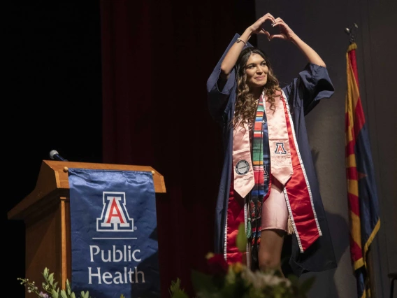 A University of Arizona Mel and Enid Zuckerman College of Public Health student makes a heart sign with her hands as she walks acroos a stage while wearing a graduation cap and gown.