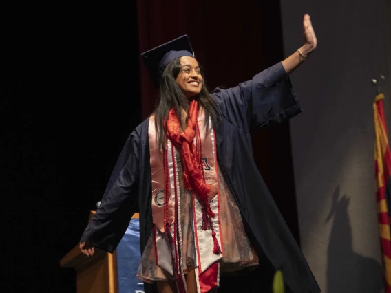 A University of Arizona Mel and Enid Zuckerman College of Public Health student, smiling and dressed in graduation regalia, waves while walking across a stage. 