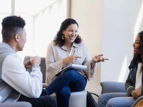 A woman with a notepad on her lap and a pen in her hand sits between two smiling college students. 