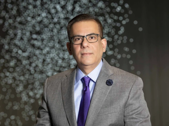 Portrait of a man wearing a grey suit, purple tie and glasses standing in front of lights in the Health Sciences Innovation Building Forum.