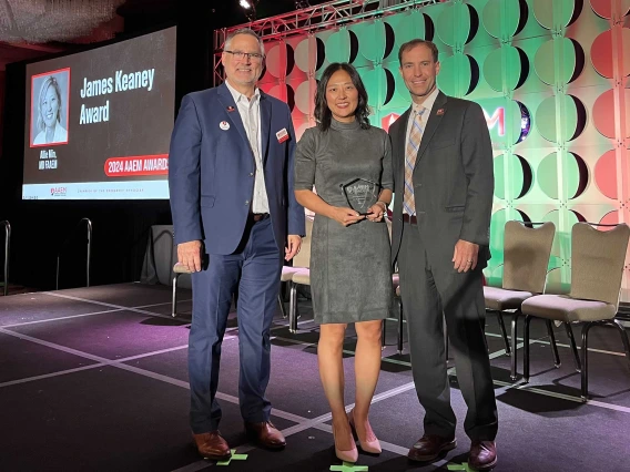 Alice Min Simpkins stands holding the James Keaney Award flanked by two male colleagues wearing suits.