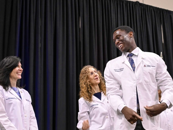 A new, male student at the University of Arizona College of Medicine – Phoenix smiles at two female professors on a stage. All are wearing medical white coats. 