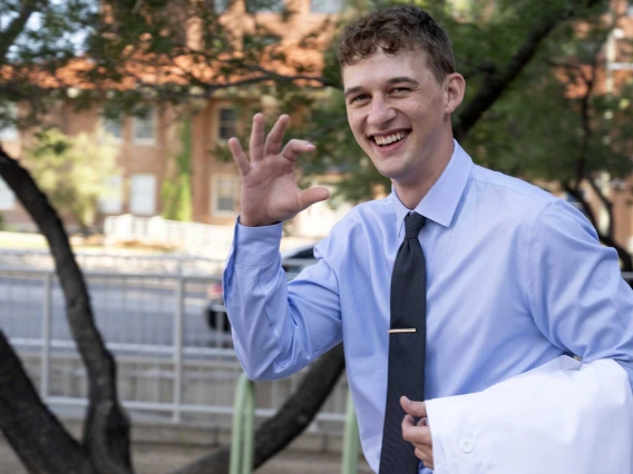 A new University of Arizona College of Medicine – Tucson student flashes the Arizona Wildcats hand sign as he walks outside with a medical white coat draped over his arm. 