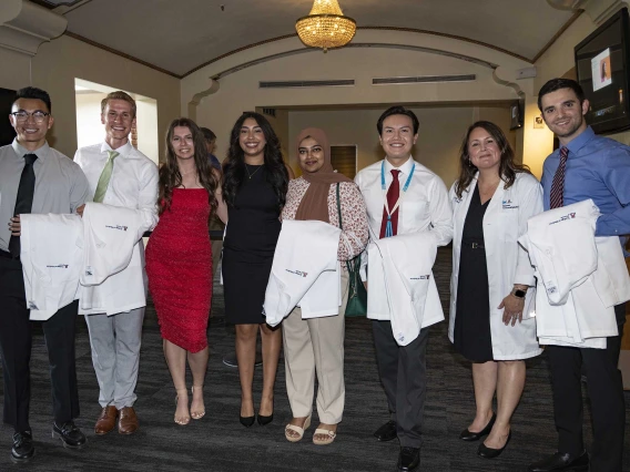 A University of Arizona College of Medicine – Tucson professor wearing a white medical coat stands with seven new medical students, all with white coats draped over their arms.