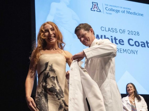 A University of Arizona College of Medicine – Tucson student in a tan print dress smiles as a professor helps her put on a white coat.