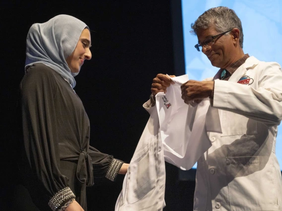 A University of Arizona College of Medicine – Tucson student wearing a black dress and gray hijab prepares to have a medical white coat put on her by a professor. 