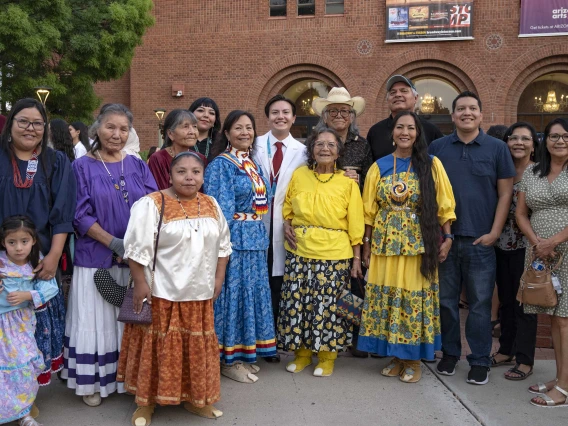 An incoming University of Arizona College of Medicine – Tucson student wearing a medical white coat stands with more than a dozen smiling family members outside near a red brick building. Several of the women in the group are wearing traditional Native American outfits.  