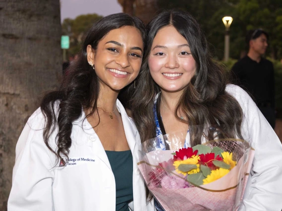 Two incoming University of Arizona College of Medicine – Tucson students wearing medical white coats stand together, smiling. One is holding flowers. 