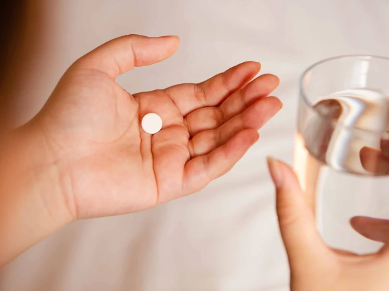 Open hand holding round white pill, other hand holding glass of water