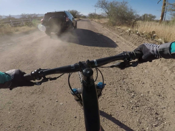 A vehicle kicks up dust as it passes a bicyclist on a dirt road