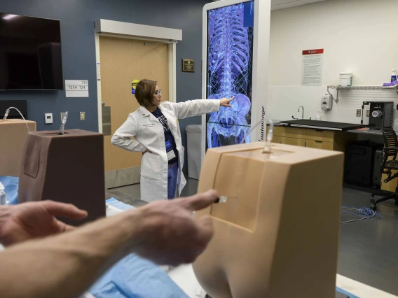 A professor in a white lab coat points to a projection of a skeleton while students in the foreground practice a procedure on the lower back of manikins. 