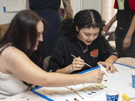 Two people sit at a table holding brushes as they paint tiles. 