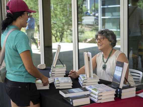 A smiling woman sits at a table covered by stacked books that she is signing while another woman stands at the table talking to her. 