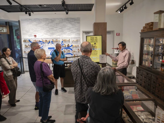 A group of about 10 people stands in the Coit Museum of Pharmacy and Health Sciences listening to Theodore Tong, PharmD, as he gives a tour. 