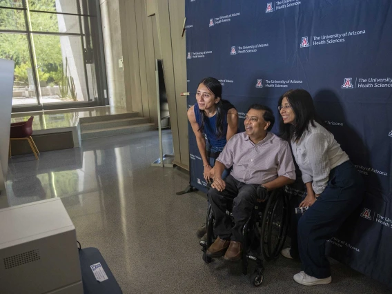 A man in a wheelchair is flanked by two women. All are smiling for a camera in front of a University of Arizona photo backdrop. 