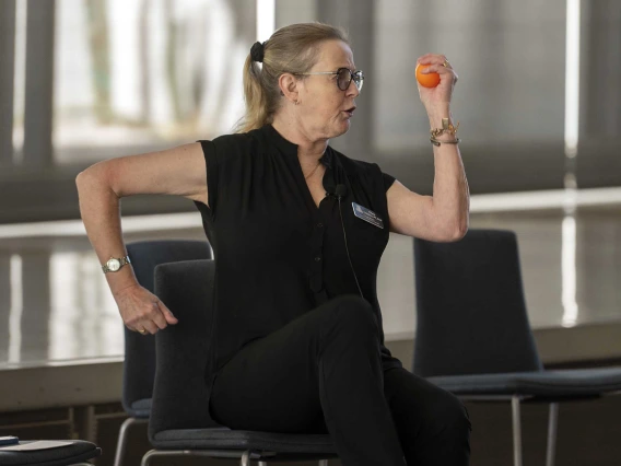 A woman sits in a chair with one arm back and the other forward, as if running in place. She holds a small ball in one hand. 