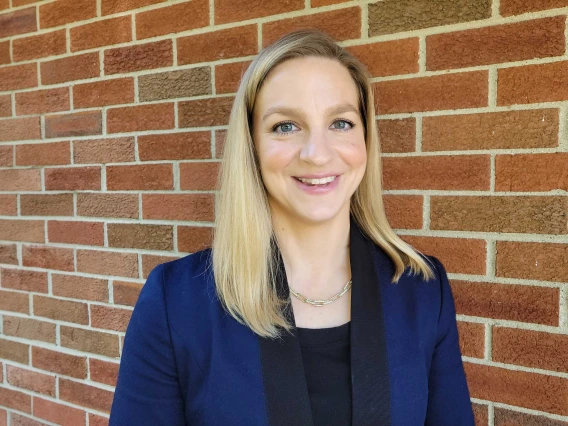 Portrait of professor Lindsay Bouchard wearing a blue jacket and smiling while standing in front of a brick wall. 