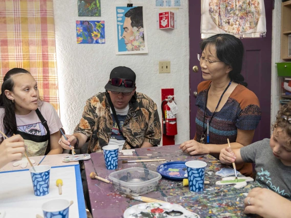 Yumi Shirai, PhD, sits at a table with a group of people who are painting at the University of Arizona’s ArtWorks program for adults with developmental disabilities. The table is covered with cups holding brushes and paints.