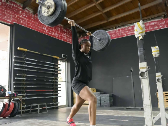 Photo of Brena Andrews in a gym holding a barbell over her head in split-jerk stance.