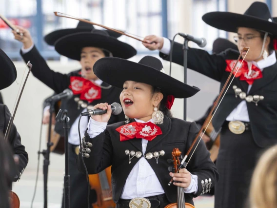 A young girl sings into a microphone in front of several other student mariachi musicians with violins who are all wearing black coats, pants and hats with red and white details. 