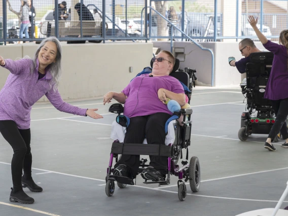 Two people using electric wheelchairs perform a dance with two people standing near them in an outdoor setting. 