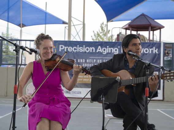 A seated woman plays a violin while a seated man plays a guitar in an outdoor settting. 