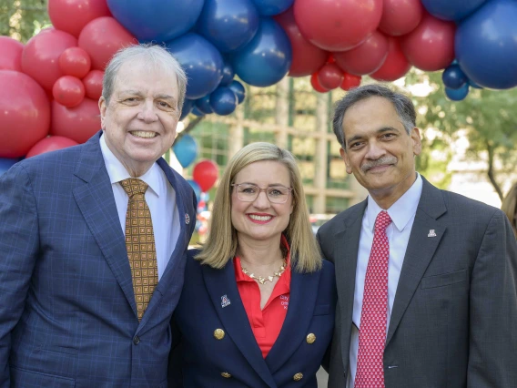 Two men in dark suits and ties stand on either side of a women in a dark blazer in an outdoor setting with red and blue balloons over their heads. All are smiling. 