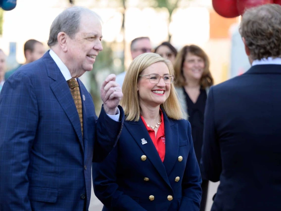 A man and woman dressed in dark blue suits stand and chat with others in an outdoor setting. Both are smiling. 