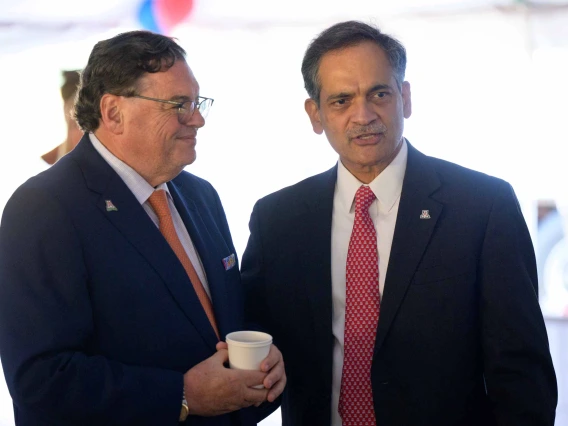 Two men in dark blue suits and red ties stand next to each other, chatting. 