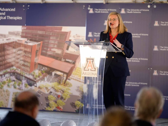 A women dressed in a dark pantsuit stands at a podium with the University of Arizona logo on it. There is a rendering of a building on a backdrop behind her. 