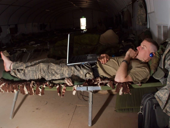 A male soldier lies on a cot while watching a laptop computer and wearing headphones.