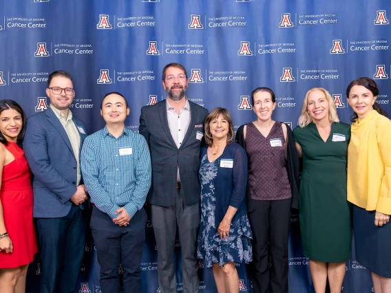 Eight members of the University of Arizona Cancer Center stand side-by-side smiling in front of a Cancer Center backdrop. 