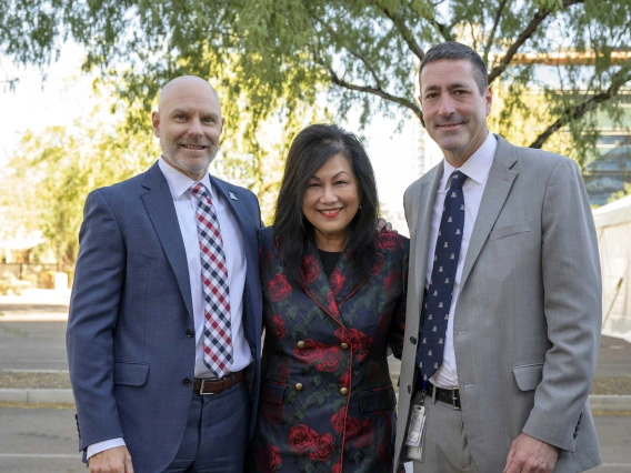 A woman flanked on either side by a man in a suit, stand outside under a tree. All three are smiling. 