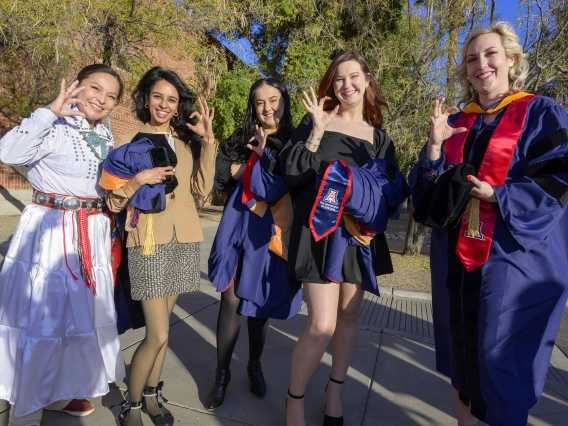 Five University of Arizona nursing students stand outside wearing graduation regalia as they smile and show the Arizona Wildcats hand sign. 