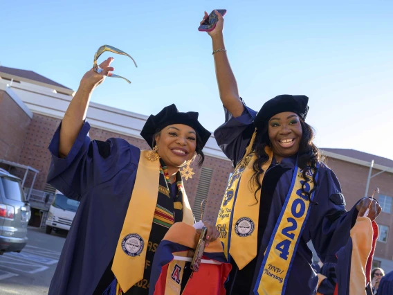 Two University of Arizona nursing students wave their arms in celebration while standing outside in their graduation caps and gowns. 