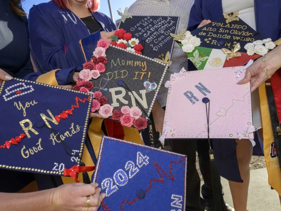 Several decorated graduation caps are held out by University of Arizona College of Nursing students. The caps have graphics and text on them noting “2024” and “RN,” along with various other messages. 