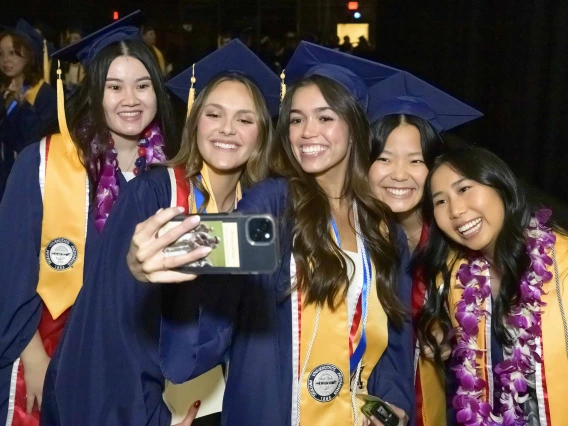 Five female nursing students wearing caps and gowns smile while taking a selfie as they stand side by side.