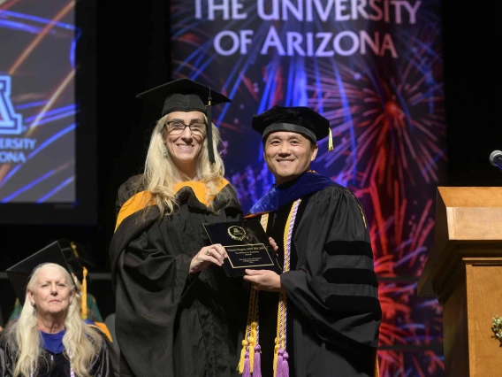 Two University of Arizona College of Nursing faculty members dressed in graduation regalia stand together on a stage, smiling and holding a plaque.  