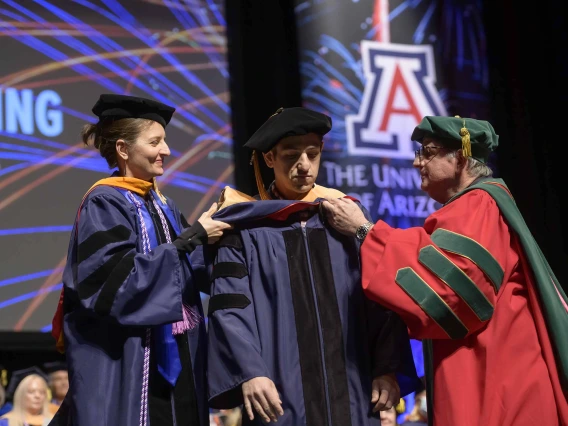 Two University of Arizona faculty members place a ceremonial hood over the shoulders of a student during a graduation ceremony. 