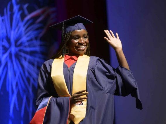 A woman in graduation regalia waves and smiles as she walks across a stage. 