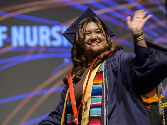 A young woman wearing graduation regalia smiles and waves as she walks across a stage. 