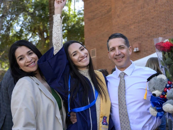 A young woman wearing a graduation gown raises her arm in celebration as she stands between her smiling parents. 